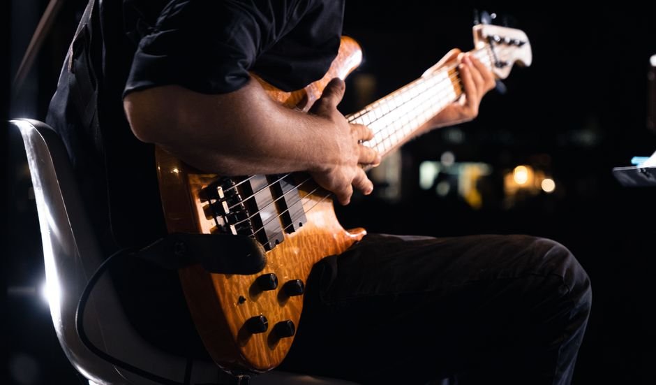 Músico sentado tocando baixo elétrico de corpo claro em palco escuro, com luzes desfocadas ao fundo