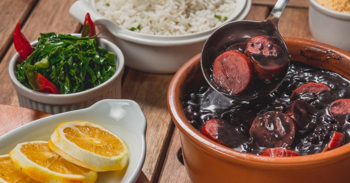 Panela de barro com feijoada ao lado de arroz, couve refogada e fatias de laranja sobre mesa de madeira