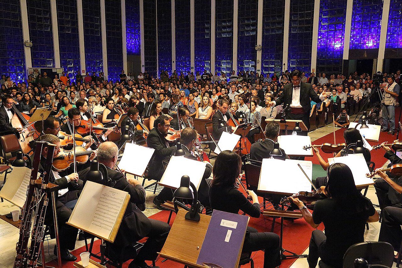 Músicos da Orquestra Sinfônica do Teatro Nacional Claudio Santoro tocando diante de plateia cheia, com o maestro de costas