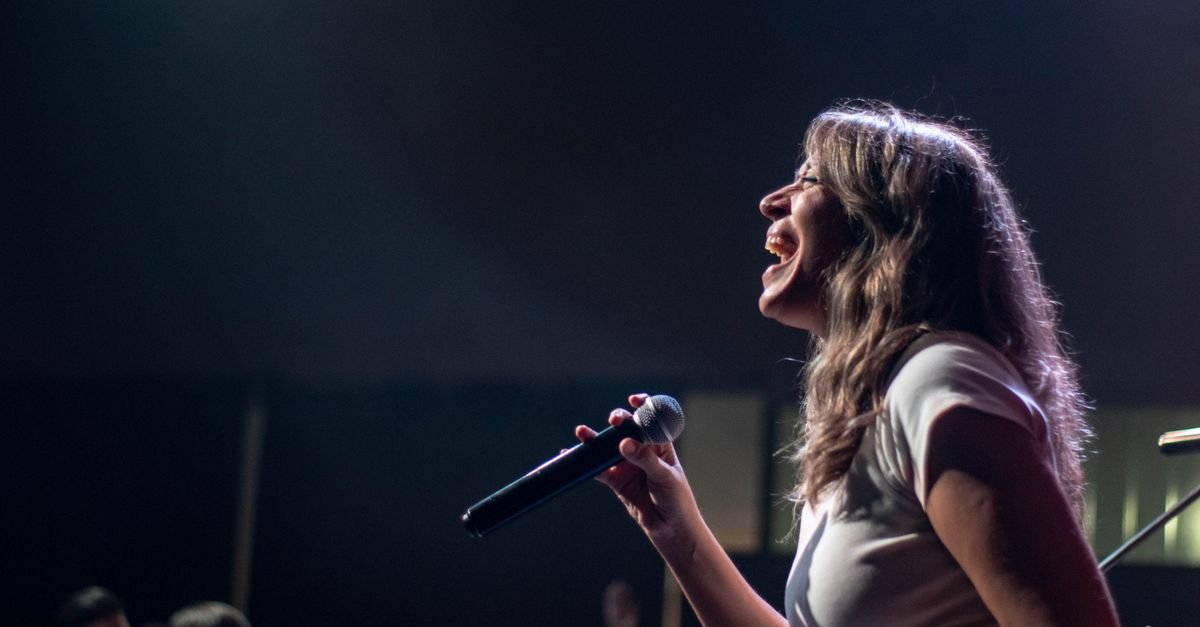 Cantora em apresentação ao vivo, de perfil, com microfone na mão sob luz de palco
