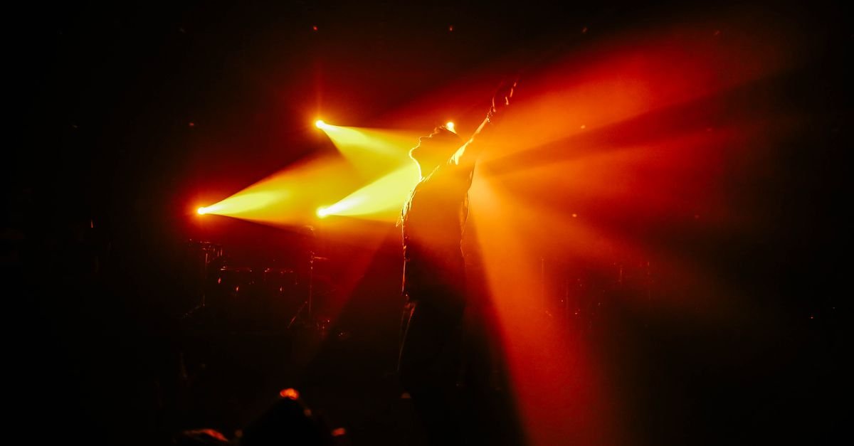 Palco de espetáculo iluminado por feixes de luz vermelha e amarela, com silhueta de artista em cena