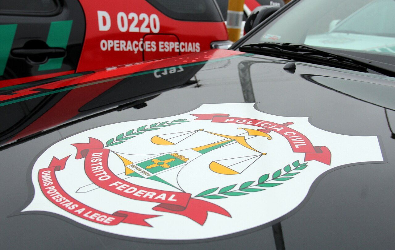 Brasão da Polícia Civil do Distrito Federal aplicado no capô de uma viatura, com carro de operações especiais ao fundo