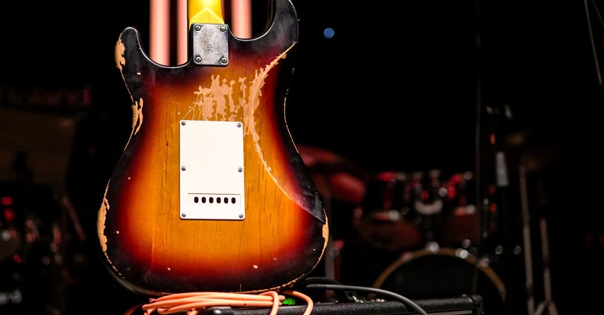 Guitarra elétrica desgastada apoiada em amplificador em palco pouco iluminado, com bateria ao fundo