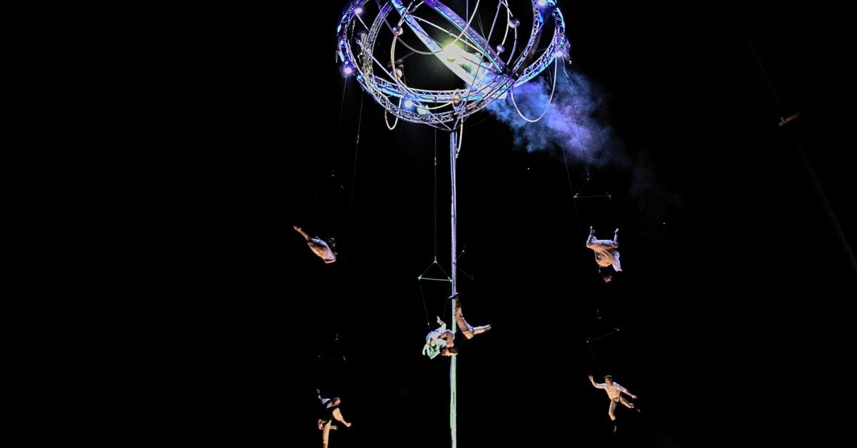 Acrobatas suspensos por cabos em apresentação aérea noturna, iluminados por refletores