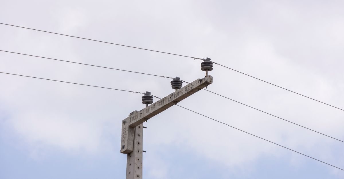 Poste de concreto com isoladores e fiação elétrica visto de baixo contra o céu claro