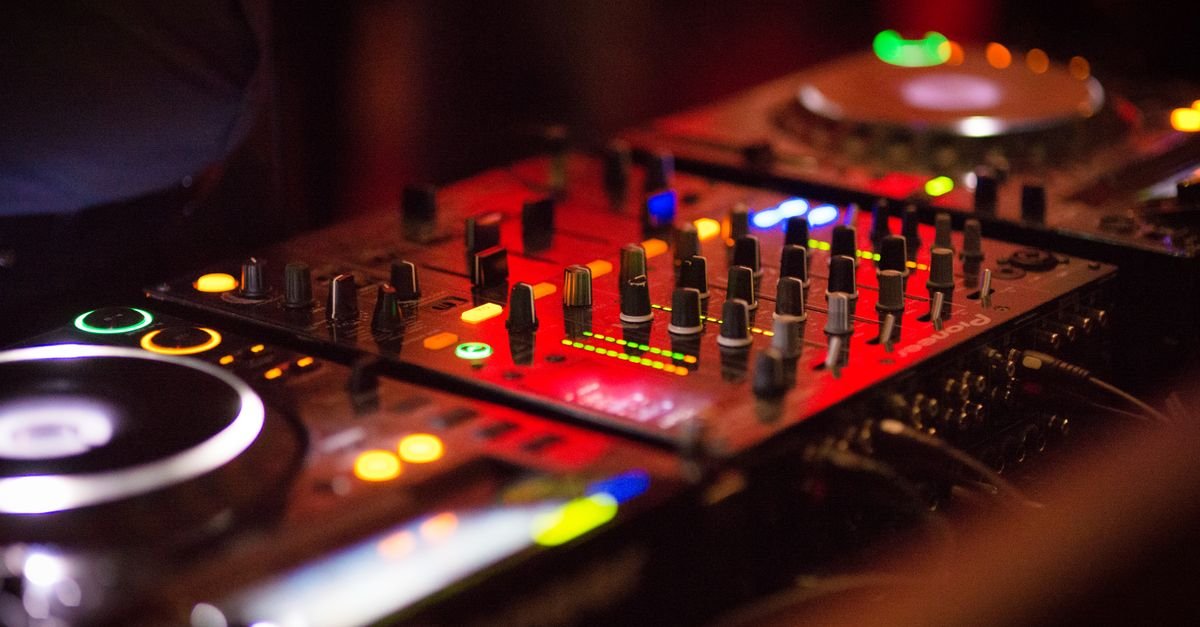 Mesa de mixagem de DJ em close, iluminada por luzes vermelhas e verdes durante uma festa