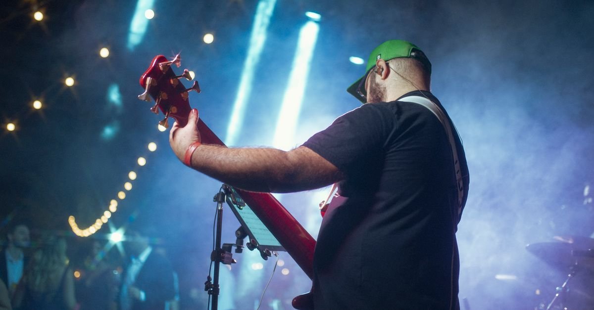 Baixista de costas tocando em palco de casa de show, com luzes azuis e fumaça ao fundo