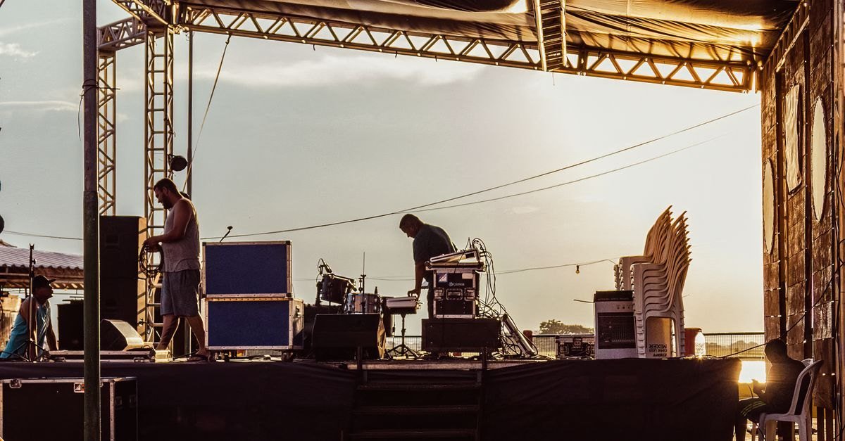 Equipe monta estrutura de palco ao ar livre durante o pôr do sol, com caixas de som e cadeiras ao redor