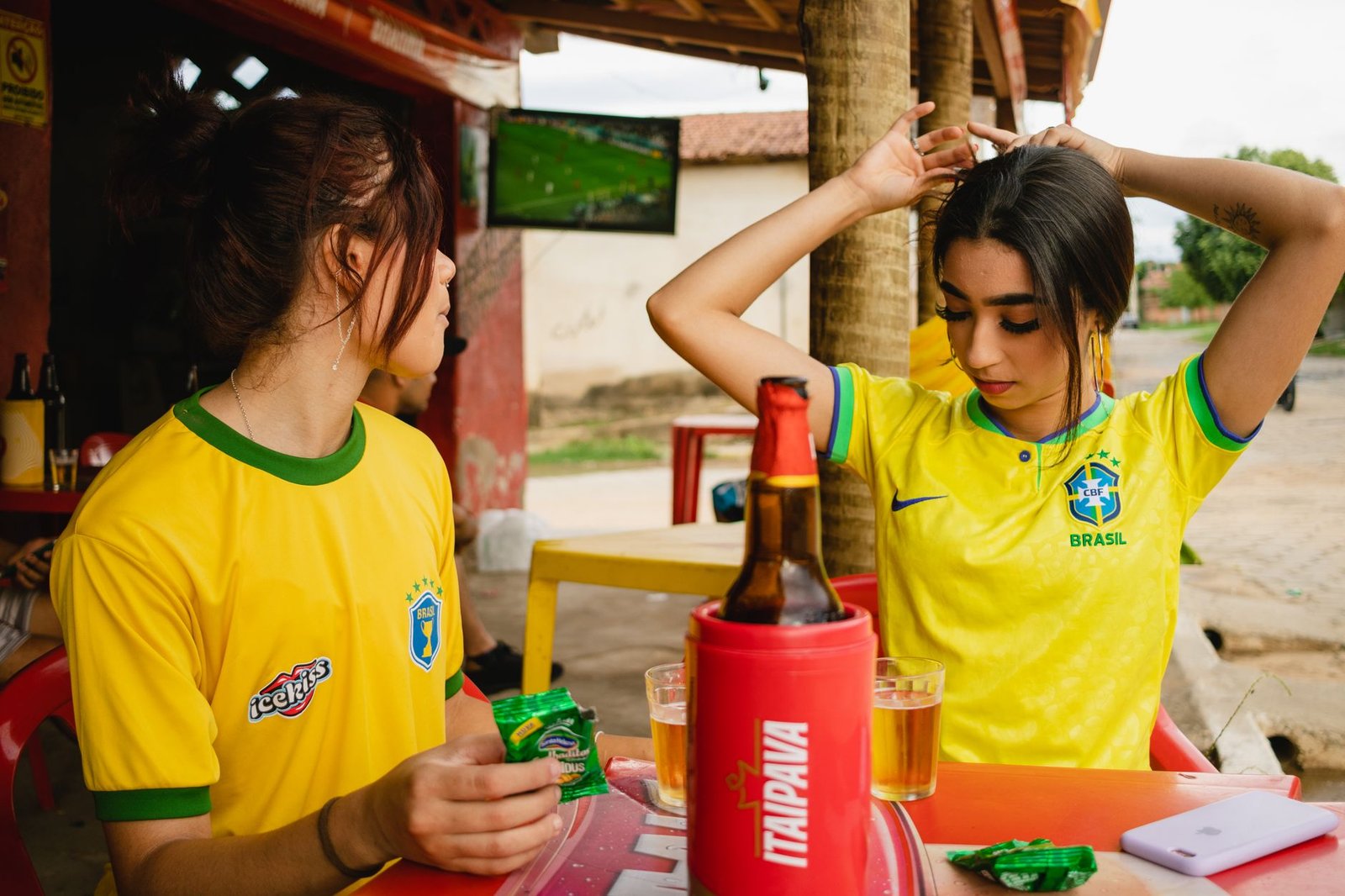 Duas amigas de camisa da seleção brasileira em mesa de bar, com TV de futebol ao fundo