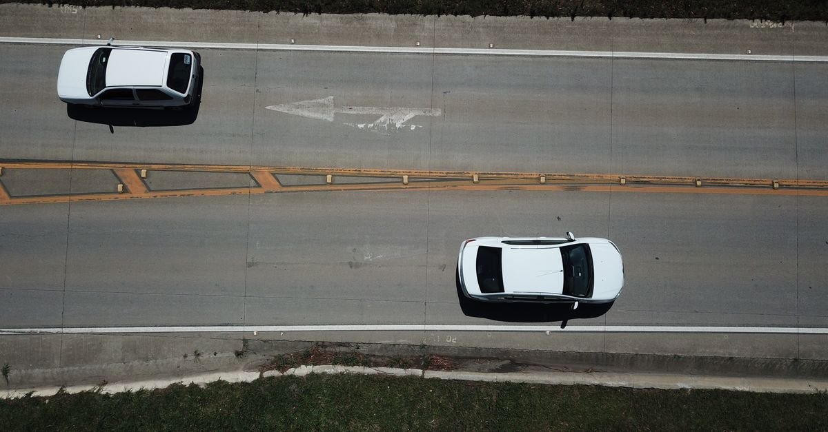 Vista aérea de cima de dois carros brancos circulando em pista asfaltada com faixa amarela central