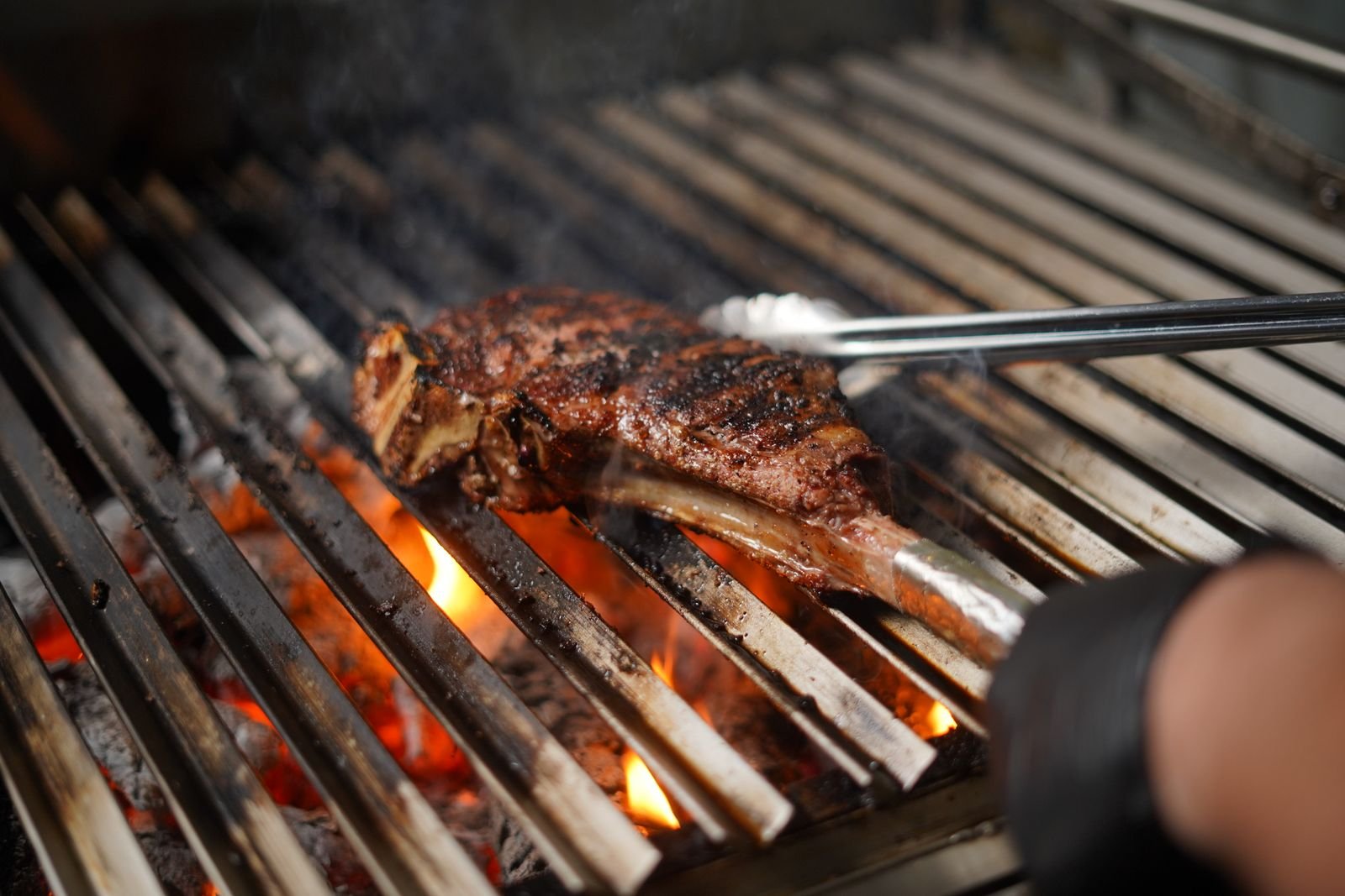 Corte de carne com osso sendo virado com pinça sobre grelha de brasa (foto ilustrativa)