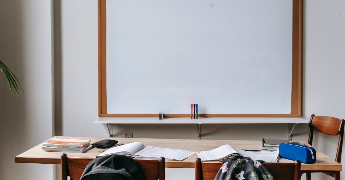 Sala de aula vazia com quadro branco, carteiras, cadernos e mochilas sobre a mesa