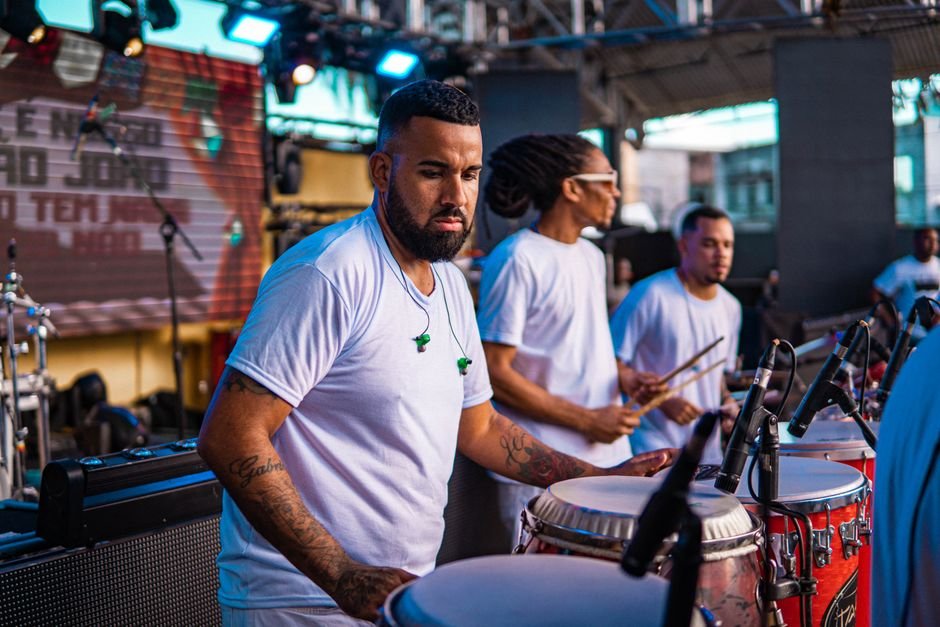 Percussionistas tocam surdo e caixa em palco ao ar livre durante apresentação de samba