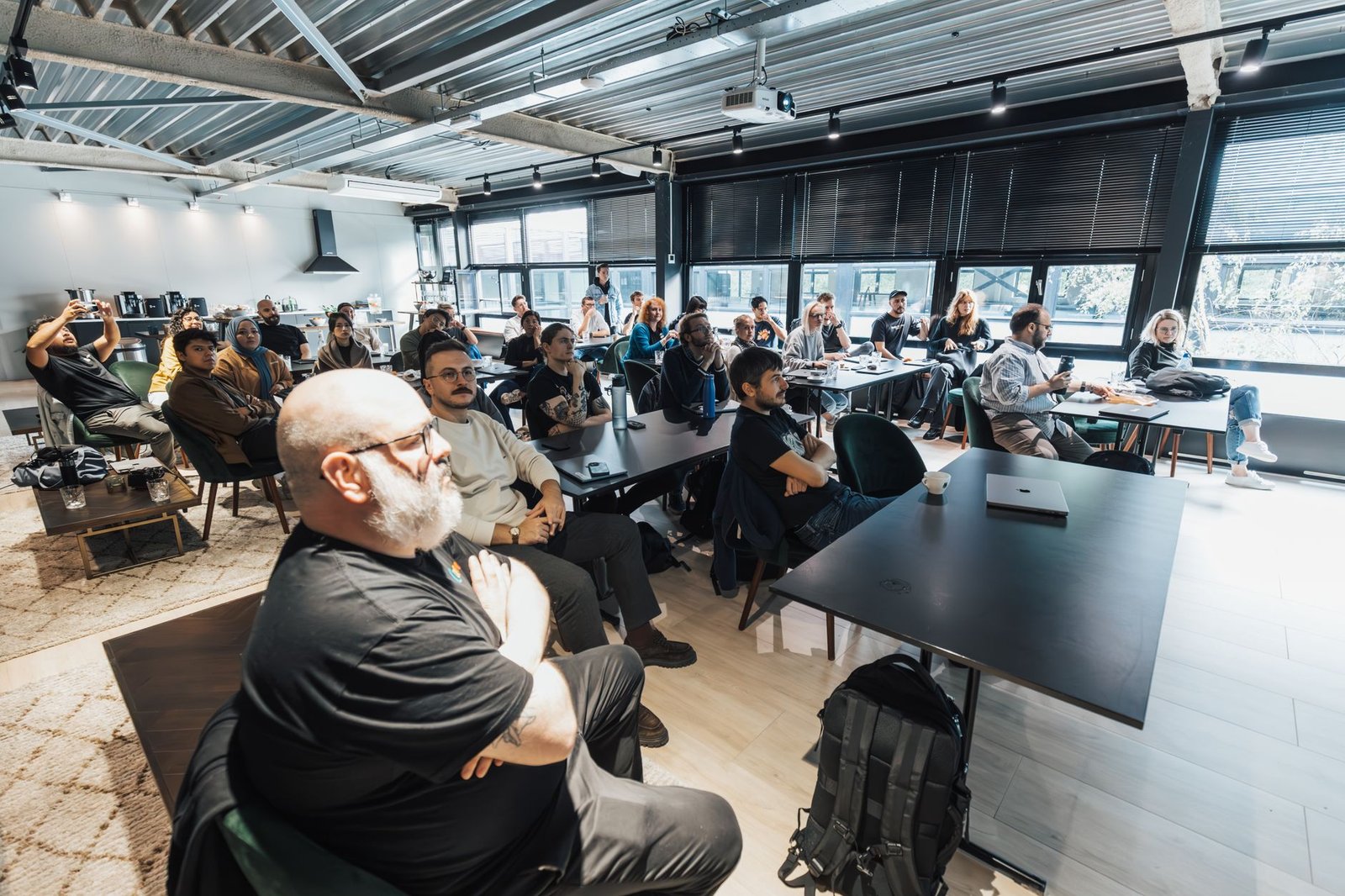 Plateia sentada em mesas e cadeiras assiste a uma apresentação em sala ampla, com projetor no teto (foto de arquivo, não é do evento)