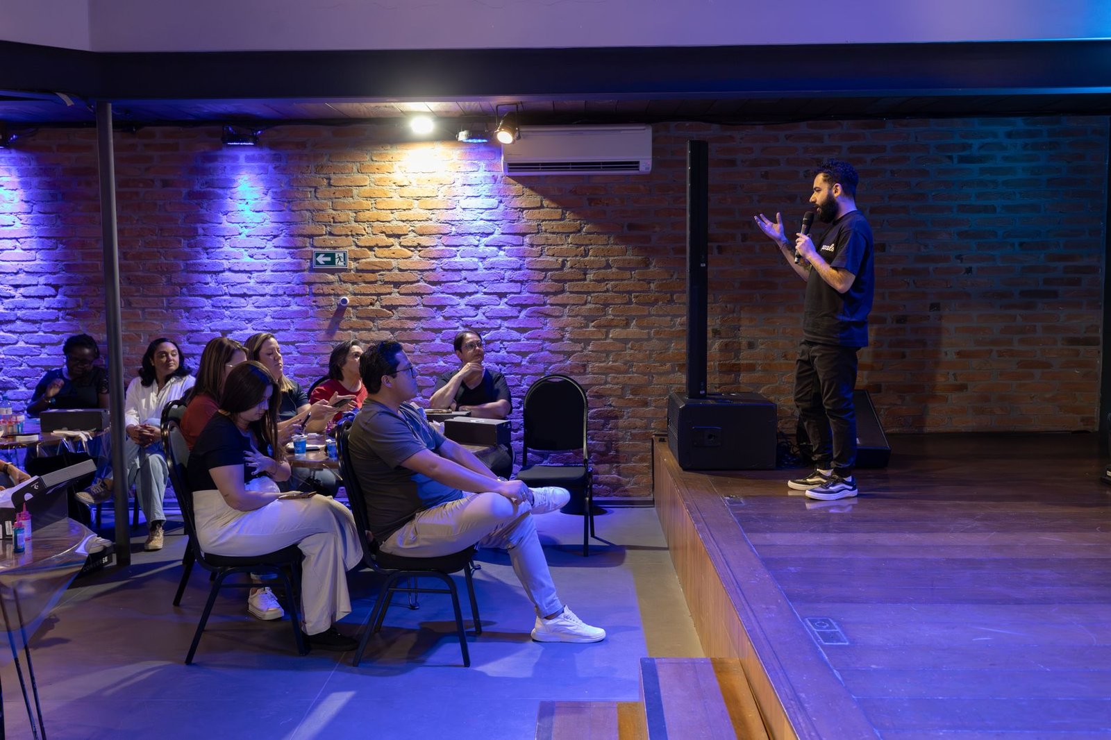 Humorista de microfone na mão se apresenta num palco baixo diante de plateia sentada, com parede de tijolos ao fundo
