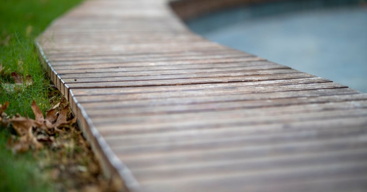 Deck de madeira contorna a borda de uma piscina, com grama ao lado