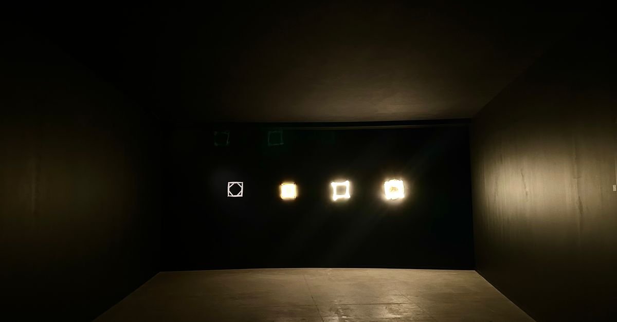 Sala escura de exposição com quatro peças luminosas alinhadas na parede ao fundo