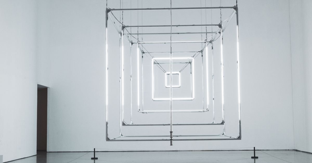 Instalação de tubos de luz em formato geométrico suspensa no centro de uma sala de galeria de paredes brancas