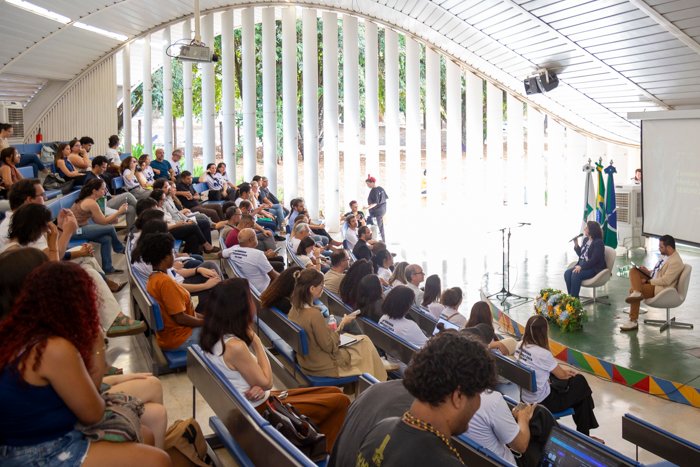 Plateia lotada no auditório do Beijódromo durante a abertura da Semana Universitária da UnB, com mesa e bandeiras ao fundo