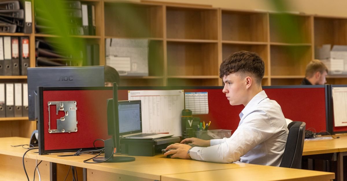 Jovem servidor trabalha diante do computador em escritório com estantes e divisórias vermelhas