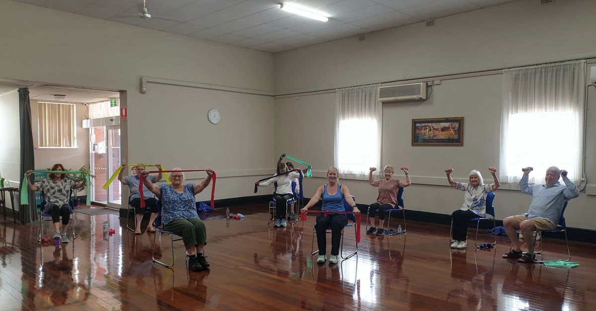 Grupo de pessoas idosas faz exercícios sentadas em cadeiras durante aula em salão