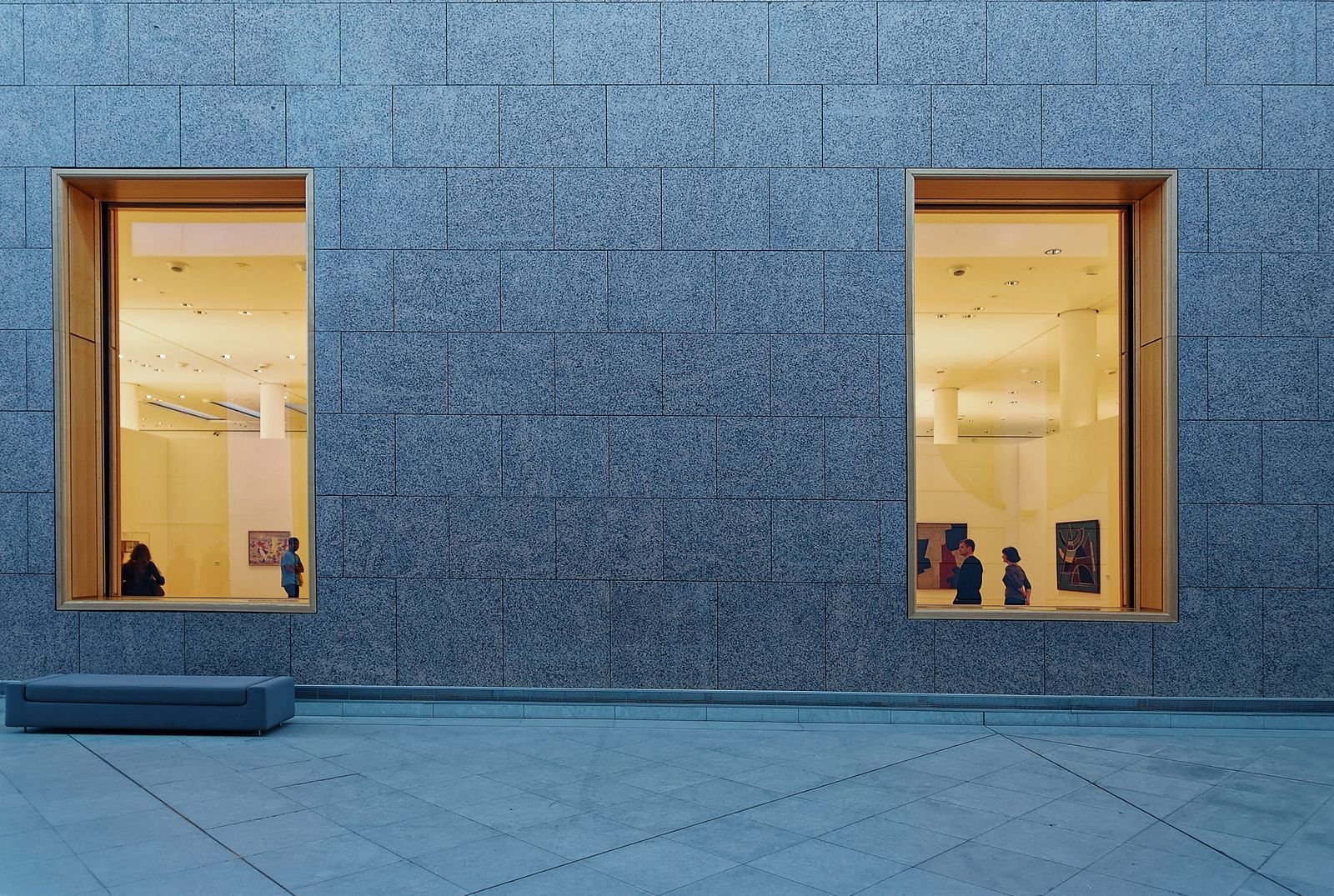 Fachada de pedra azulada de um museu ao entardecer, com duas janelas amplas iluminadas mostrando visitantes na galeria.