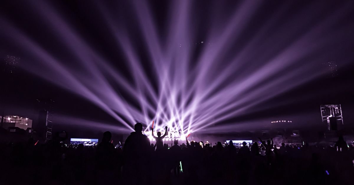Plateia em silhueta diante de palco iluminado por feixes de luz durante show noturno