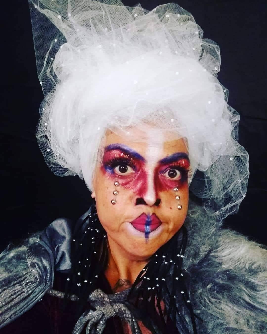 Artista com maquiagem cênica colorida, peruca branca e véu de tule encara a câmera