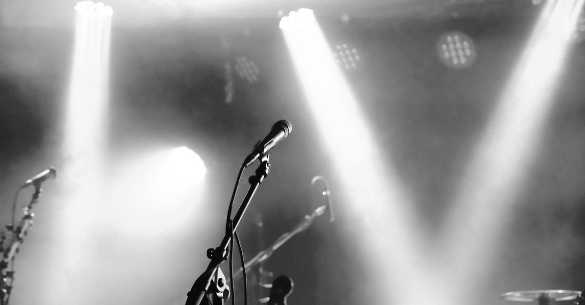 Microfone em pedestal no centro de um palco iluminado por refletores, em preto e branco