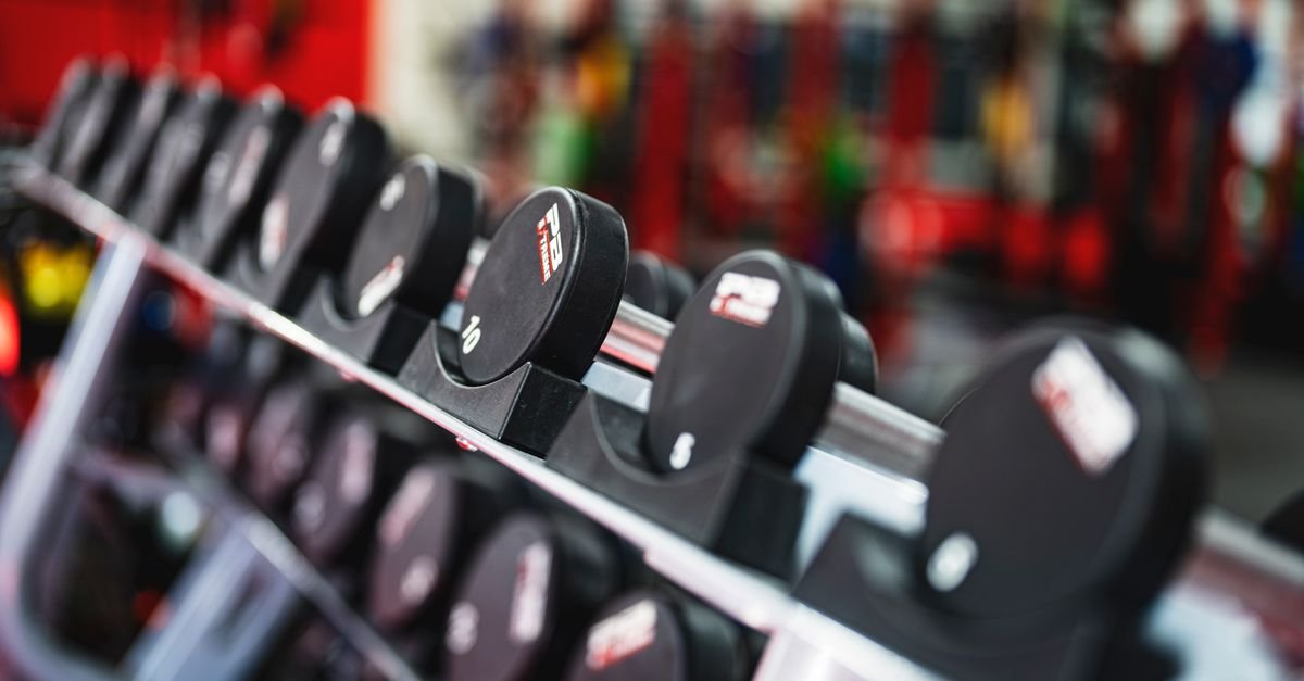 Halteres pretos enfileirados em suporte de academia, com o ambiente desfocado ao fundo