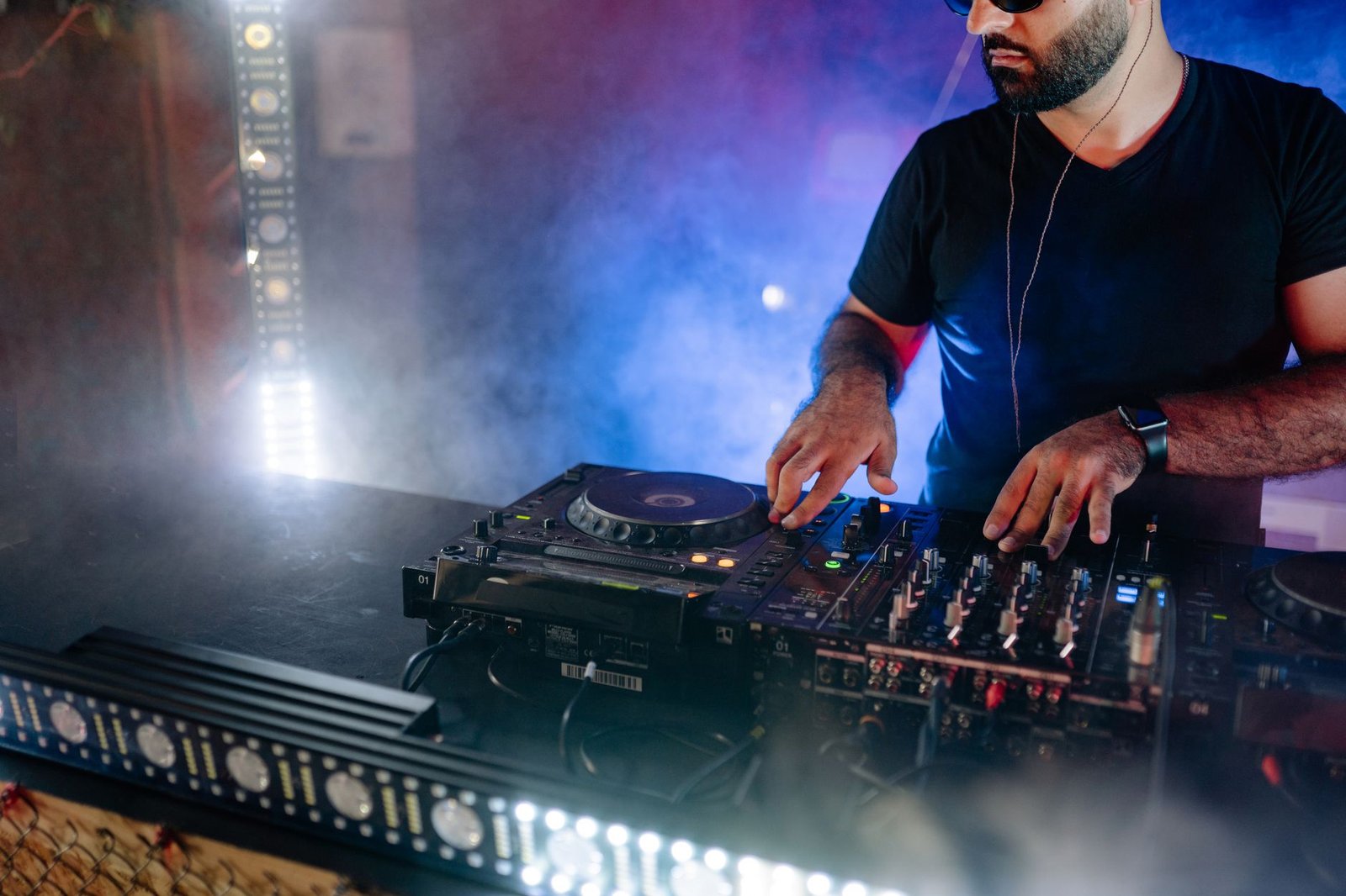 DJ de óculos escuros opera controladora e toca-discos em cabine com fumaça e luz azul