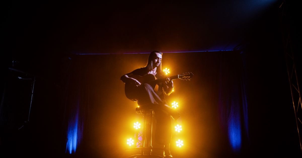 Músico toca violão sentado em banco alto sob luz amarela no palco