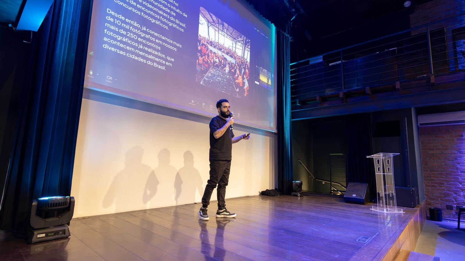 Palestrante em pé no palco de teatro com telão ao fundo durante apresentação