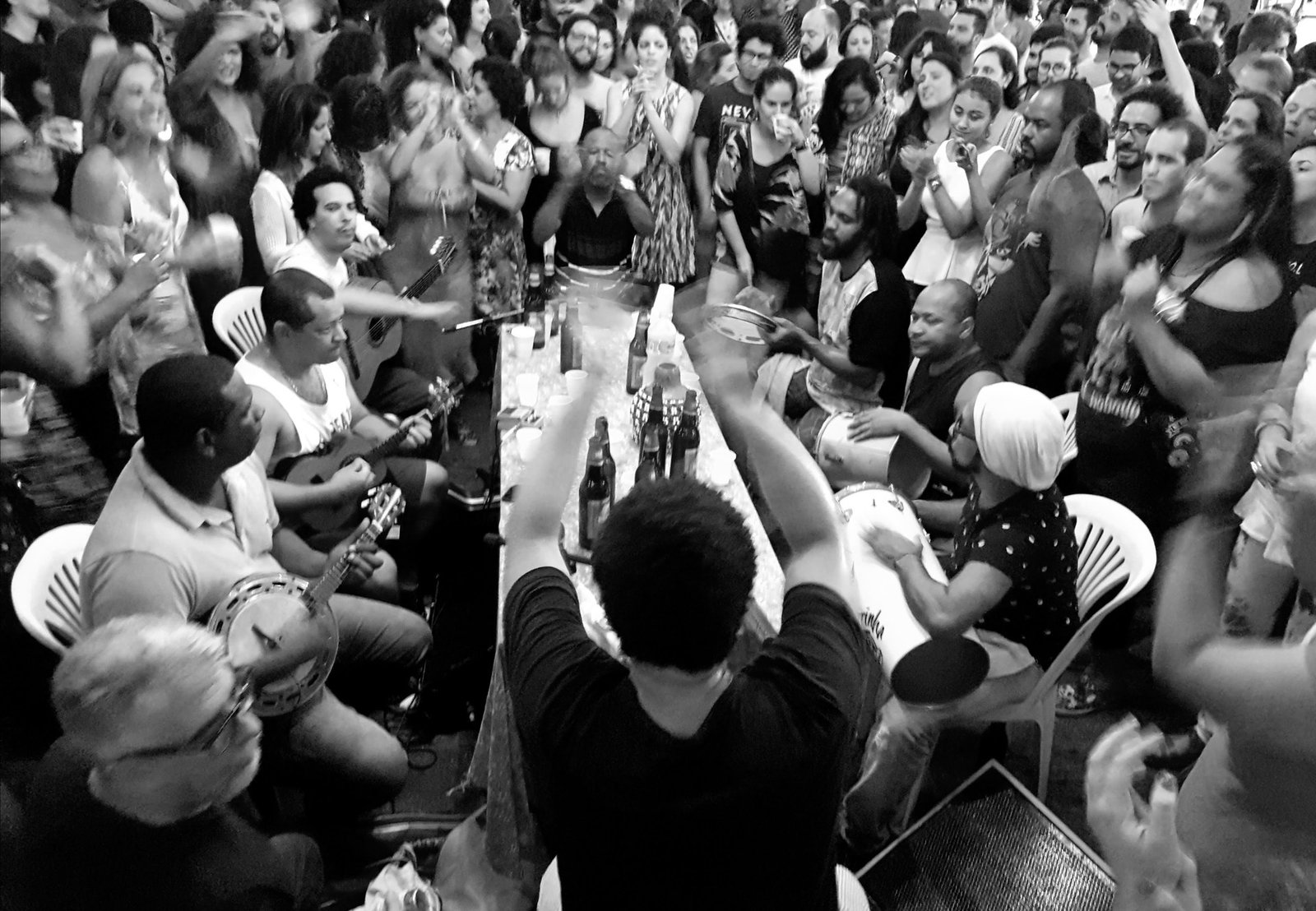 Roda de samba lotada em preto e branco, com violão, cavaquinho, banjo e percussão ao redor de uma mesa (foto ilustrativa de uma roda no Rio de Janeiro)