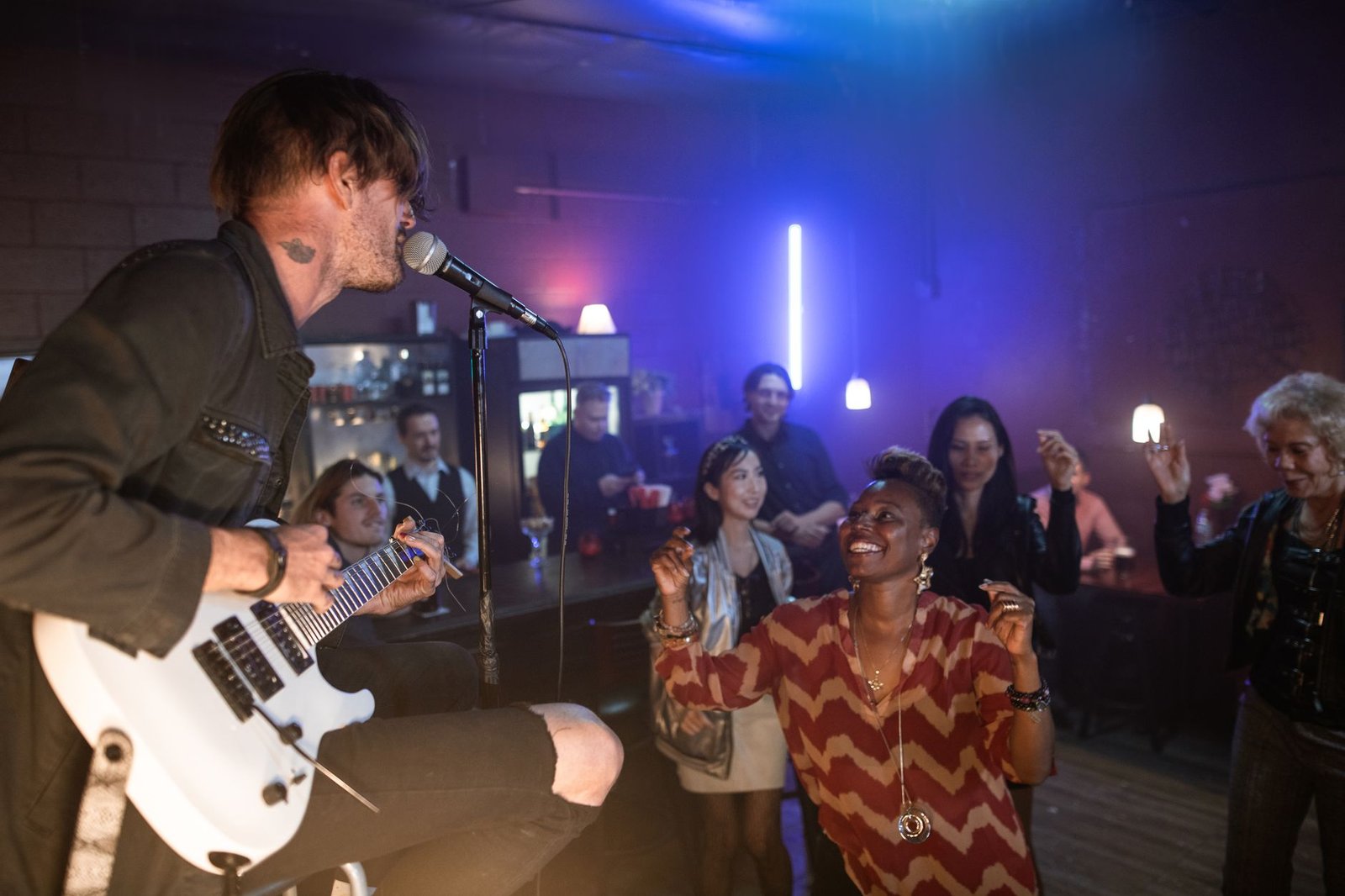 Cantor com guitarra em bar de música ao vivo, com pessoas dançando ao fundo