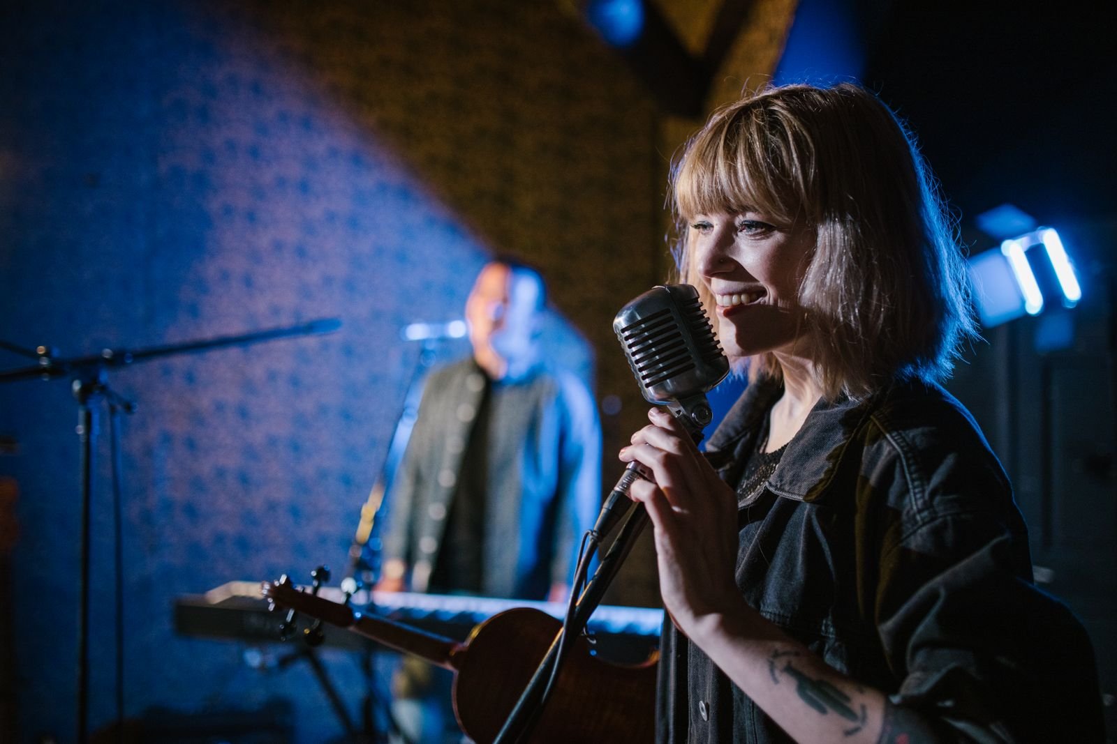Cantora sorri diante de um microfone antigo em palco com luz azul, com músico ao teclado ao fundo (foto ilustrativa)