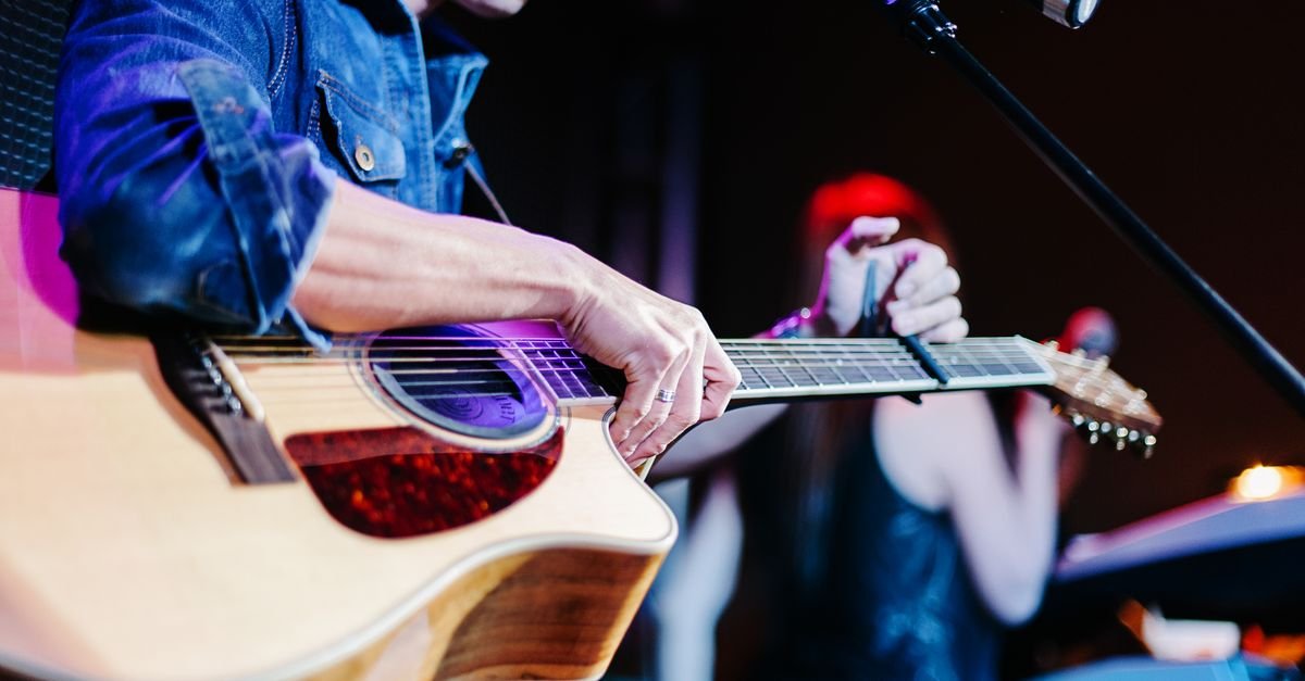 Close de músico tocando violão em palco de show, com luz colorida ao fundo