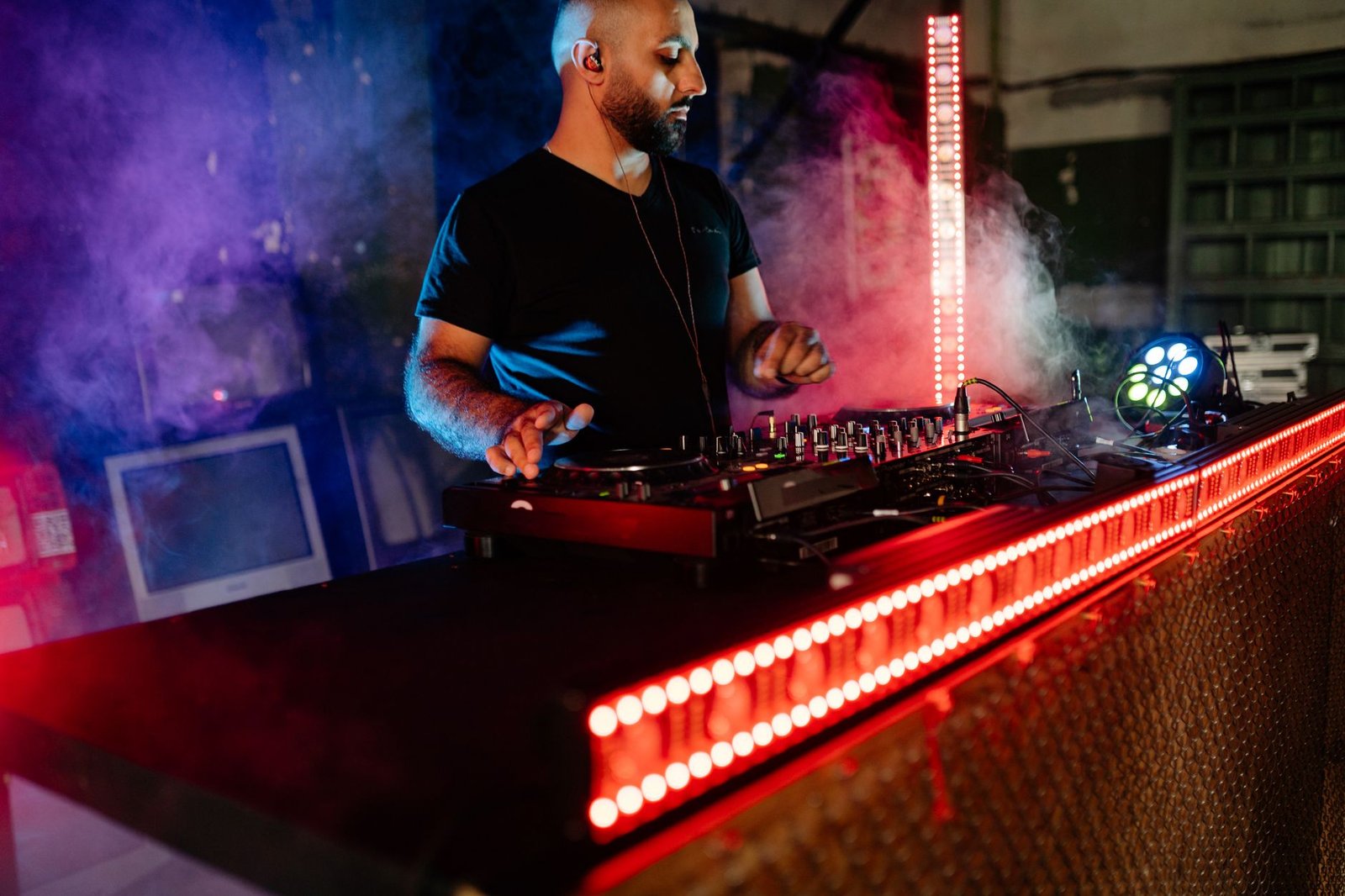 DJ de camiseta preta e fone de ouvido opera mesa de som sob luzes vermelhas e fumaça (foto ilustrativa)
