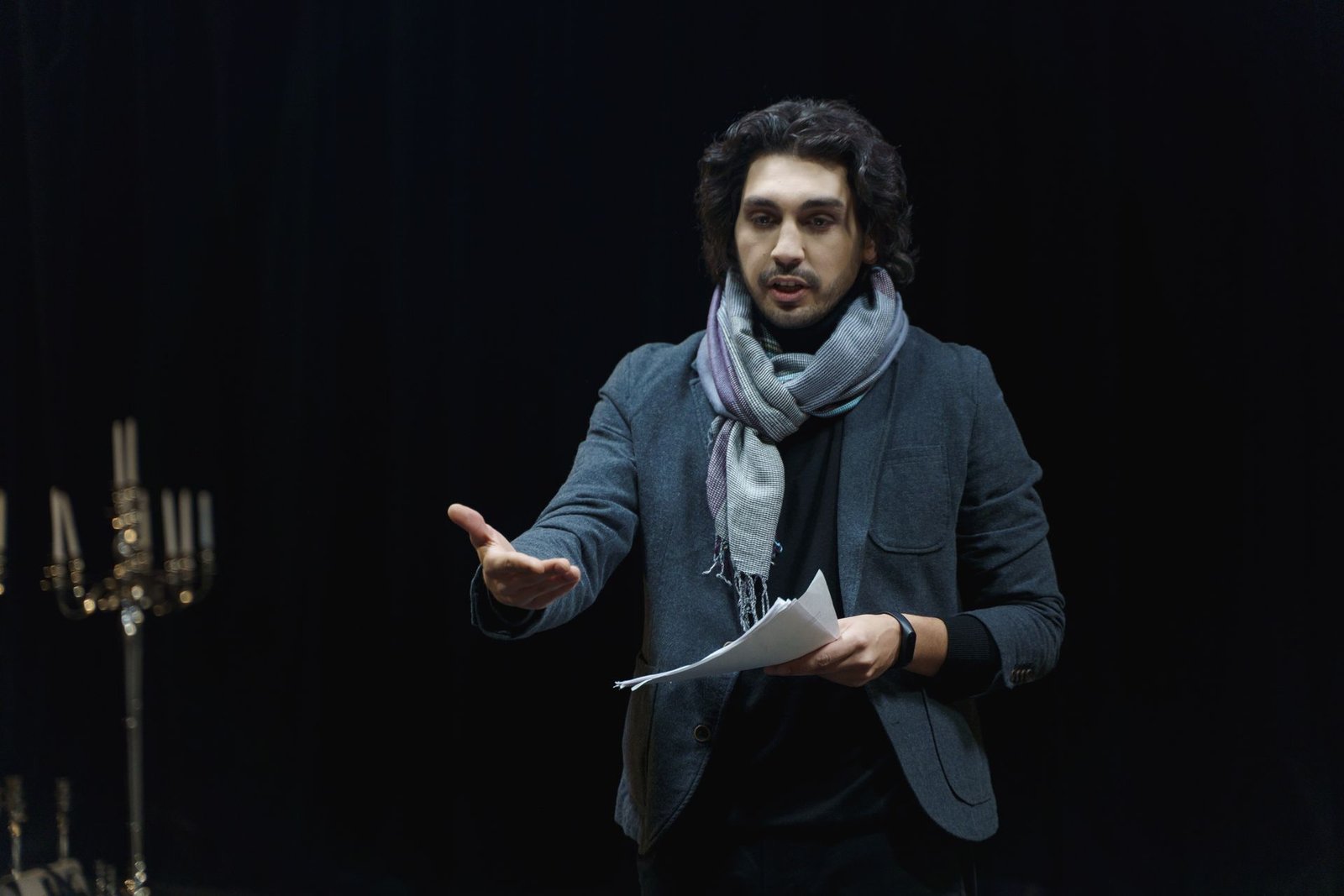 Ator gesticula sobre um palco escuro com o roteiro na mão