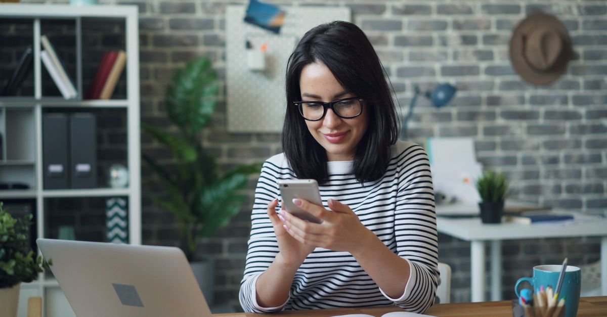 Mulher sorri enquanto usa o celular em uma mesa de escritório com notebook aberto