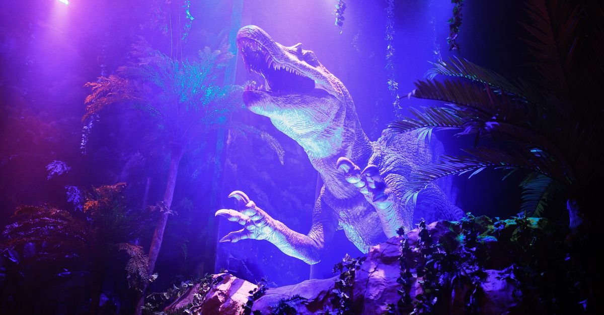 Dinossauro animatrônico de grande porte em cenário de floresta iluminado por luz azul e roxa