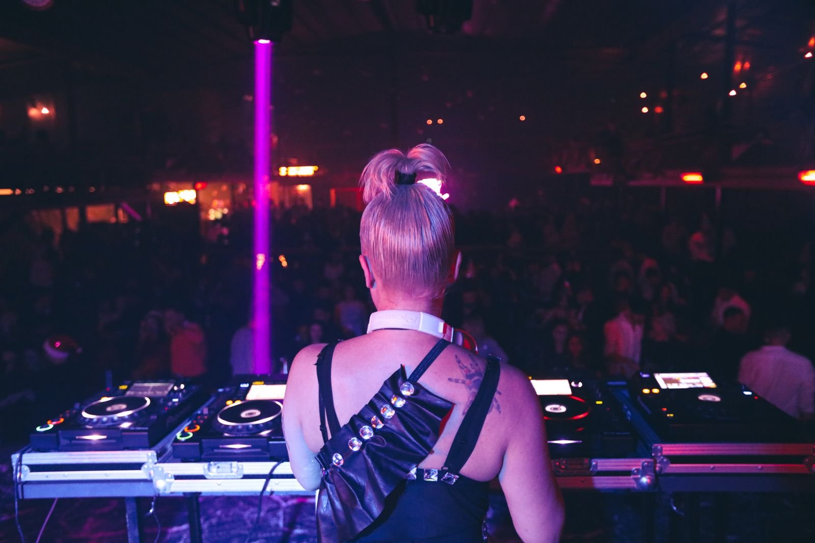 DJ de costas diante dos CDJs, com luz roxa e pista cheia ao fundo, em foto de arquivo de casa noturna