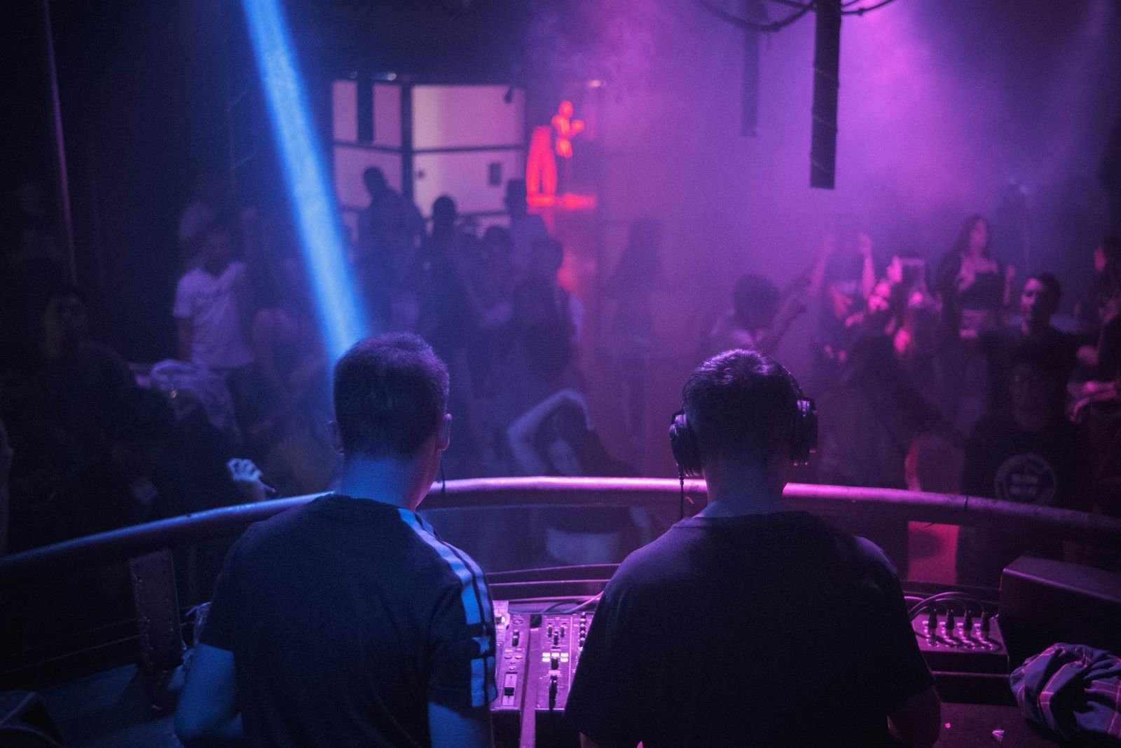 Dois DJs de costas diante da pista de uma casa noturna, com luz azul e fumaça roxa
