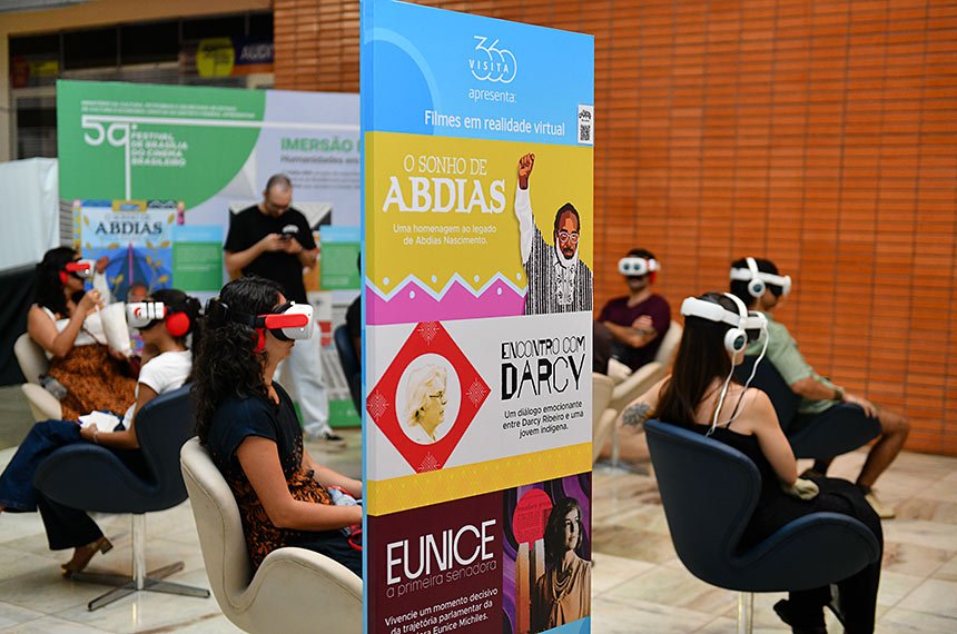Público sentado em poltronas giratórias usa óculos de realidade virtual diante de banner do projeto Visita 360