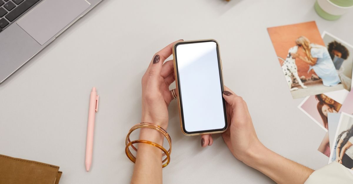Mãos de uma mulher segurando um smartphone com a tela ligada, sobre uma mesa clara