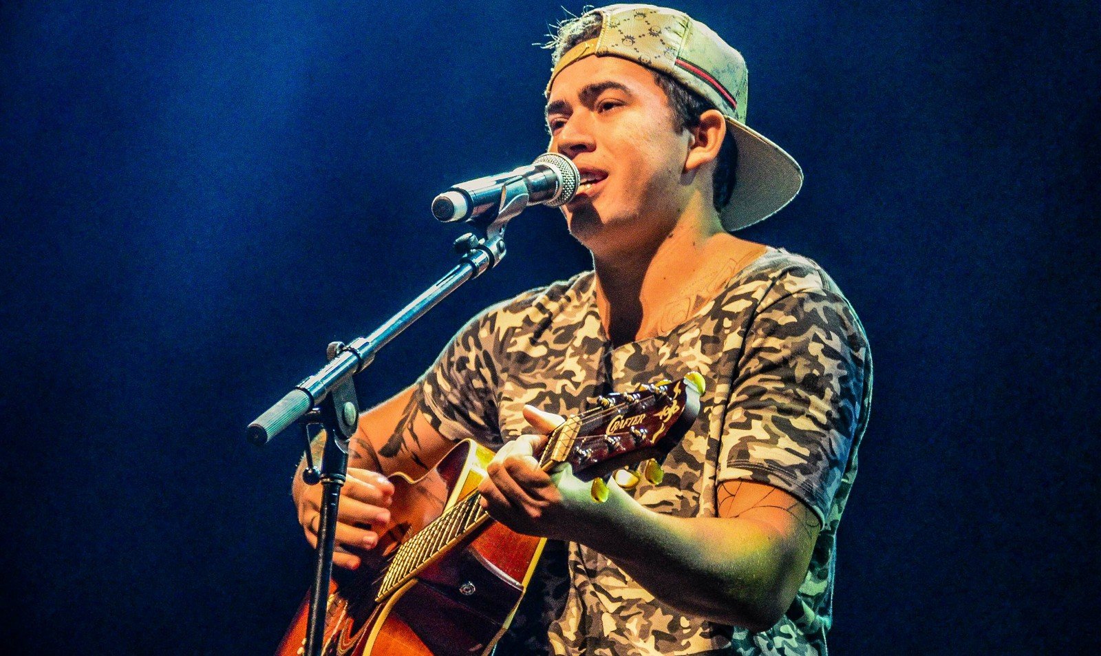 Whindersson Nunes canta ao microfone com violão durante show, sob luz azul de palco