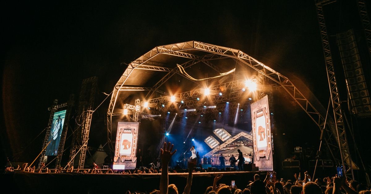 Palco de show ao ar livre iluminado à noite, com público de braços erguidos